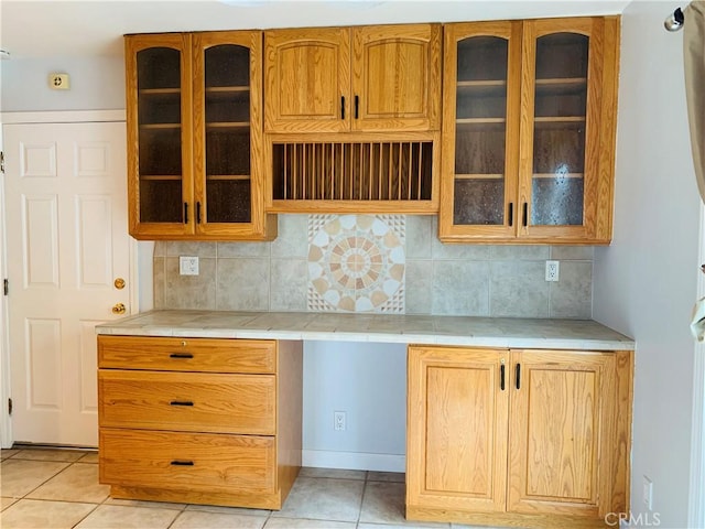 kitchen featuring backsplash, glass insert cabinets, tile counters, light tile patterned floors, and built in desk