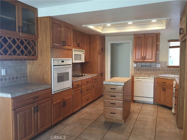 kitchen featuring white appliances, tile countertops, light tile patterned floors, a raised ceiling, and backsplash