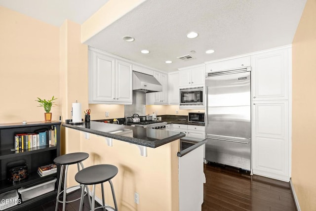 kitchen with visible vents, white cabinets, built in appliances, a peninsula, and wall chimney range hood