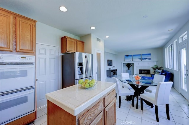 kitchen with tile counters, recessed lighting, double oven, a glass covered fireplace, and stainless steel fridge