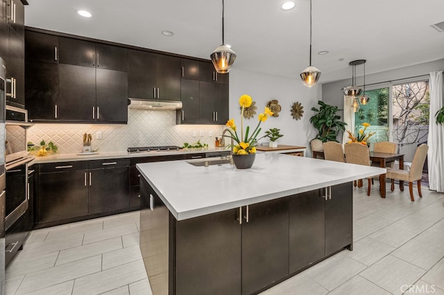 kitchen with under cabinet range hood, a sink, light countertops, a center island with sink, and pendant lighting