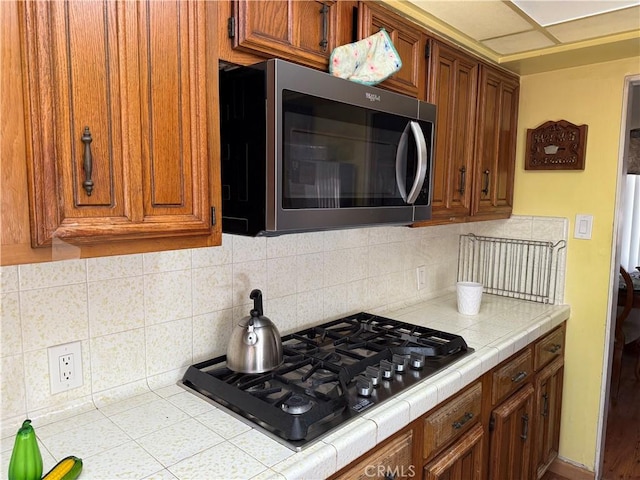 kitchen featuring black gas stovetop, backsplash, tile counters, brown cabinets, and stainless steel microwave