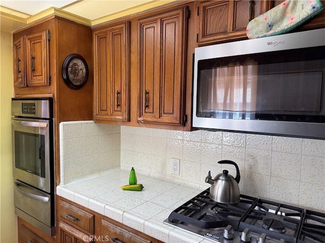 kitchen featuring a warming drawer, tile countertops, stainless steel microwave, brown cabinetry, and gas cooktop