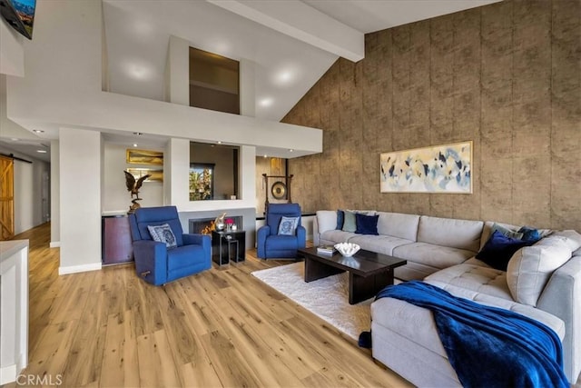 living area with a warm lit fireplace, high vaulted ceiling, a barn door, light wood-style floors, and beam ceiling