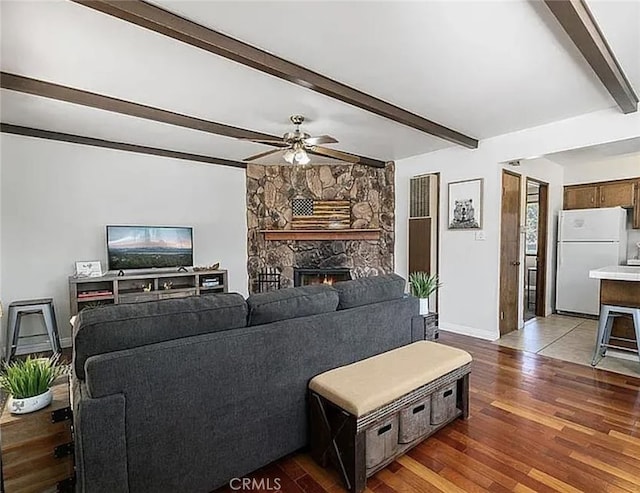 living room with a stone fireplace, a ceiling fan, baseboards, beam ceiling, and dark wood-style floors