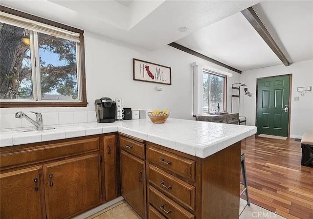 kitchen featuring tile counters, a peninsula, a sink, light wood-type flooring, and beam ceiling