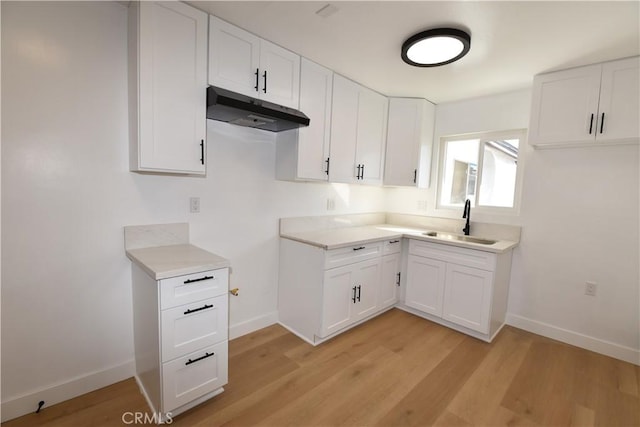 kitchen featuring light wood finished floors, baseboards, white cabinets, under cabinet range hood, and a sink