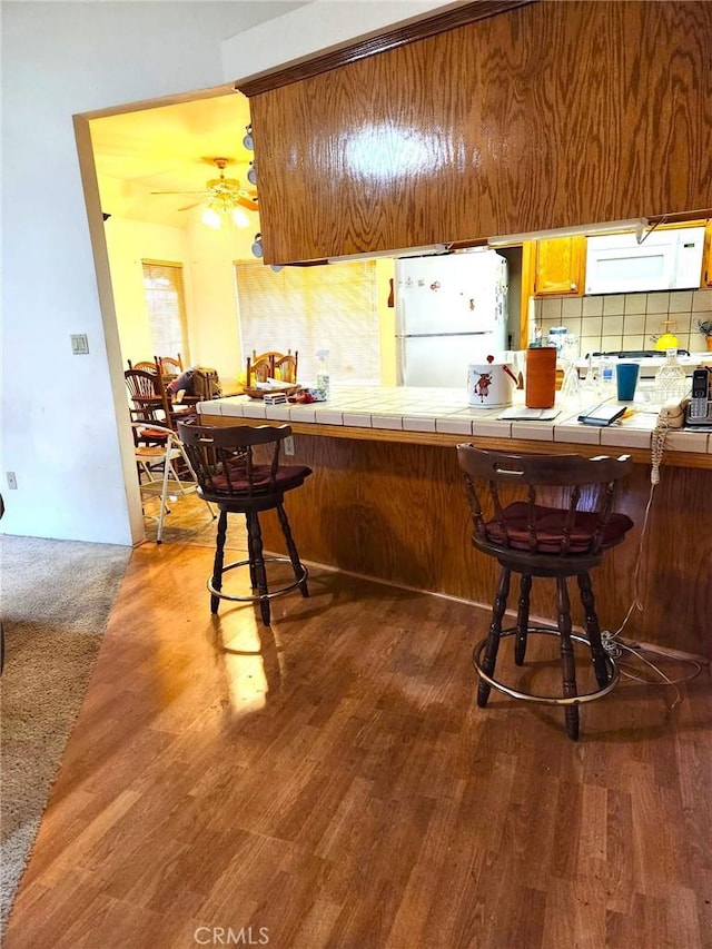 kitchen with a breakfast bar area, white appliances, backsplash, tile counters, and brown cabinets