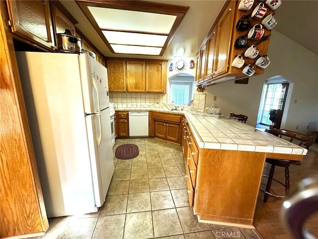 kitchen with arched walkways, a breakfast bar, brown cabinets, white appliances, and a peninsula