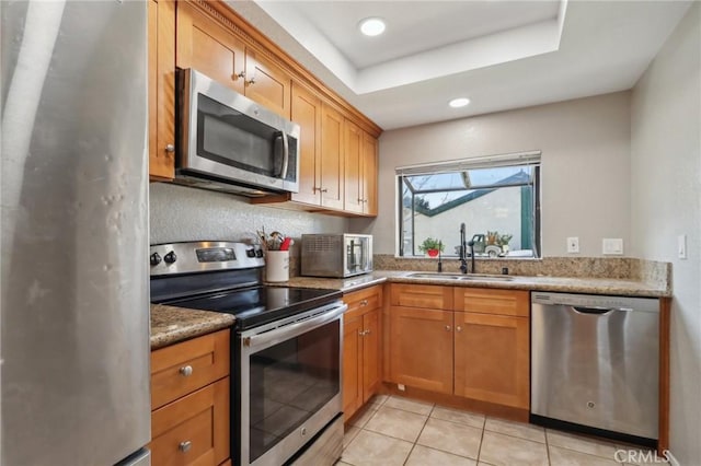 kitchen with brown cabinetry, light tile patterned flooring, stainless steel appliances, a raised ceiling, and a sink