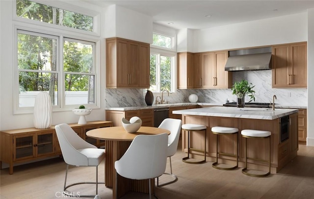 kitchen with light wood-style flooring, wall chimney exhaust hood, a center island with sink, dishwasher, and tasteful backsplash
