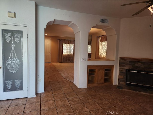 unfurnished living room featuring visible vents, arched walkways, a ceiling fan, tile patterned flooring, and a stone fireplace