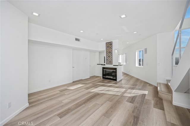 unfurnished living room with visible vents, baseboards, a glass covered fireplace, light wood-style flooring, and stairs