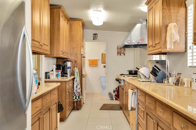 kitchen featuring light tile patterned floors, brown cabinetry, wall chimney exhaust hood, freestanding refrigerator, and light countertops