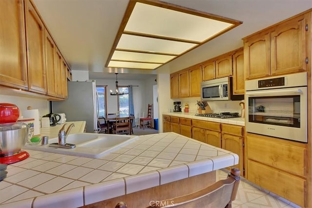 kitchen featuring tile counters, brown cabinets, stainless steel appliances, pendant lighting, and a sink