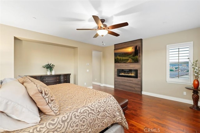 bedroom featuring a large fireplace, ceiling fan, baseboards, and wood finished floors