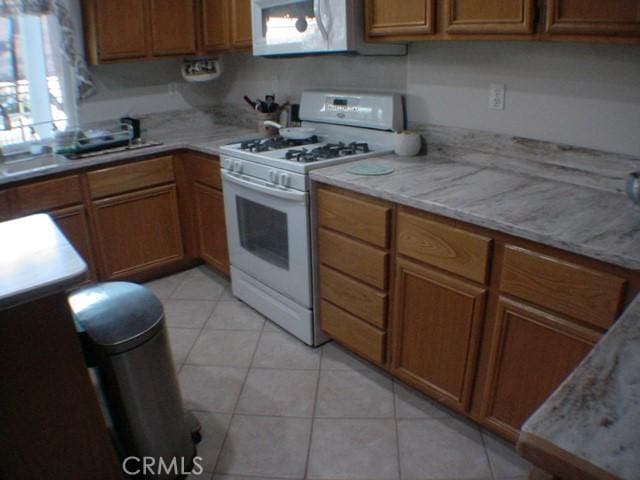 kitchen featuring brown cabinetry, white appliances, light tile patterned flooring, and light stone countertops