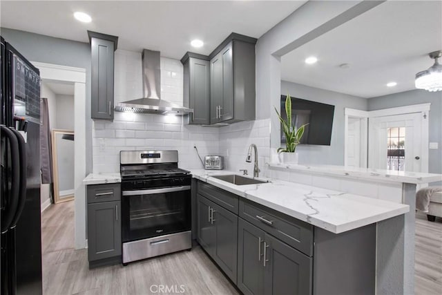 kitchen with stainless steel range with gas cooktop, a peninsula, gray cabinets, a sink, and wall chimney exhaust hood