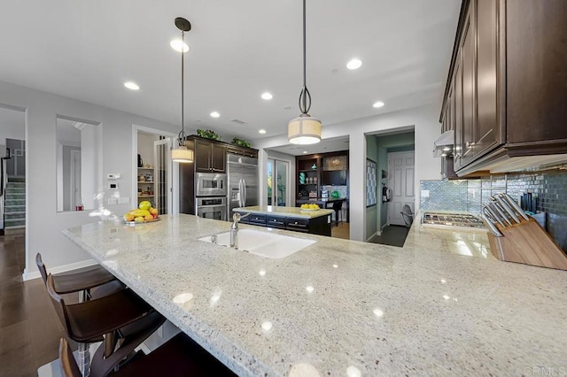 kitchen with light stone counters, built in appliances, a peninsula, pendant lighting, and a sink