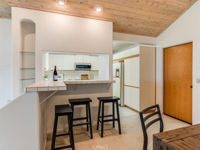 kitchen with white refrigerator with ice dispenser, tile counters, stainless steel microwave, wood ceiling, and white cabinetry