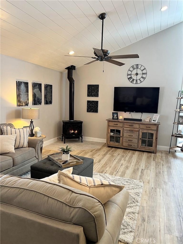 living room featuring a ceiling fan, a wood stove, vaulted ceiling, light wood-type flooring, and baseboards