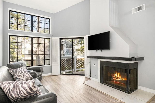 living room featuring light wood finished floors, visible vents, baseboards, a lit fireplace, and a high ceiling