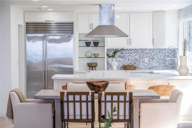 kitchen with stainless steel built in fridge, island exhaust hood, light countertops, white cabinetry, and backsplash