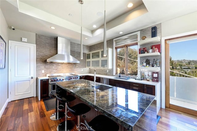 kitchen with dark stone counters, wall chimney exhaust hood, a tray ceiling, high end stove, and a kitchen bar