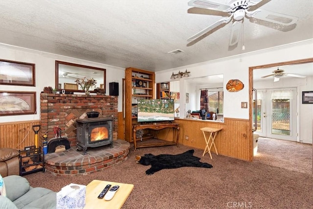 carpeted living room featuring wooden walls, visible vents, wainscoting, a wood stove, and a textured ceiling