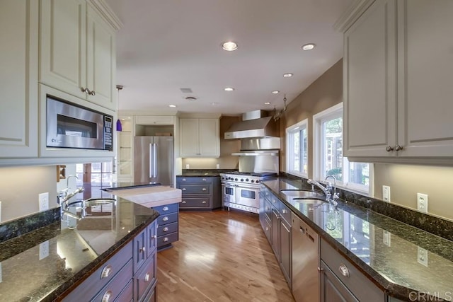 kitchen featuring appliances with stainless steel finishes, white cabinetry, a sink, dark stone counters, and wall chimney exhaust hood