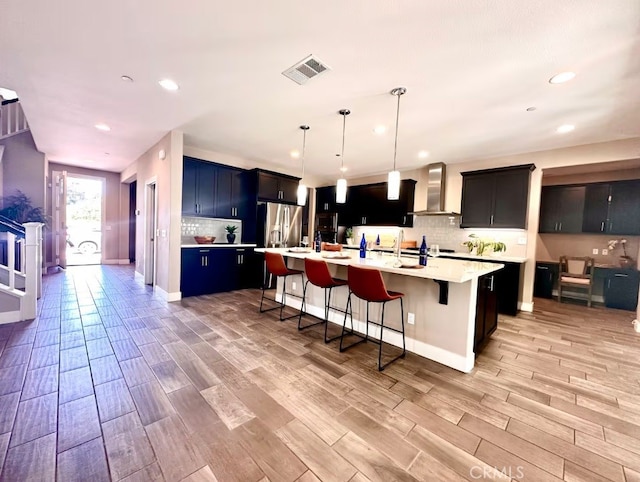 kitchen with visible vents, light countertops, a large island with sink, wall chimney range hood, and pendant lighting