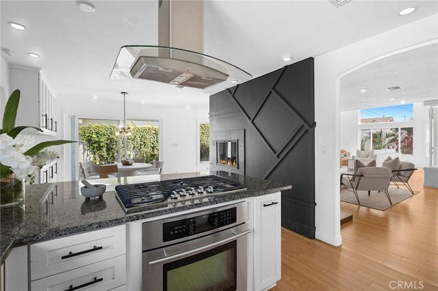 kitchen with island exhaust hood, stainless steel appliances, a multi sided fireplace, and white cabinets