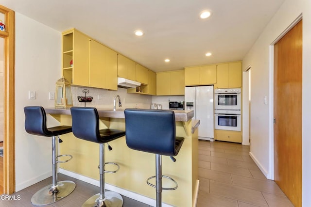 kitchen with white refrigerator with ice dispenser, open shelves, double oven, a peninsula, and a kitchen breakfast bar