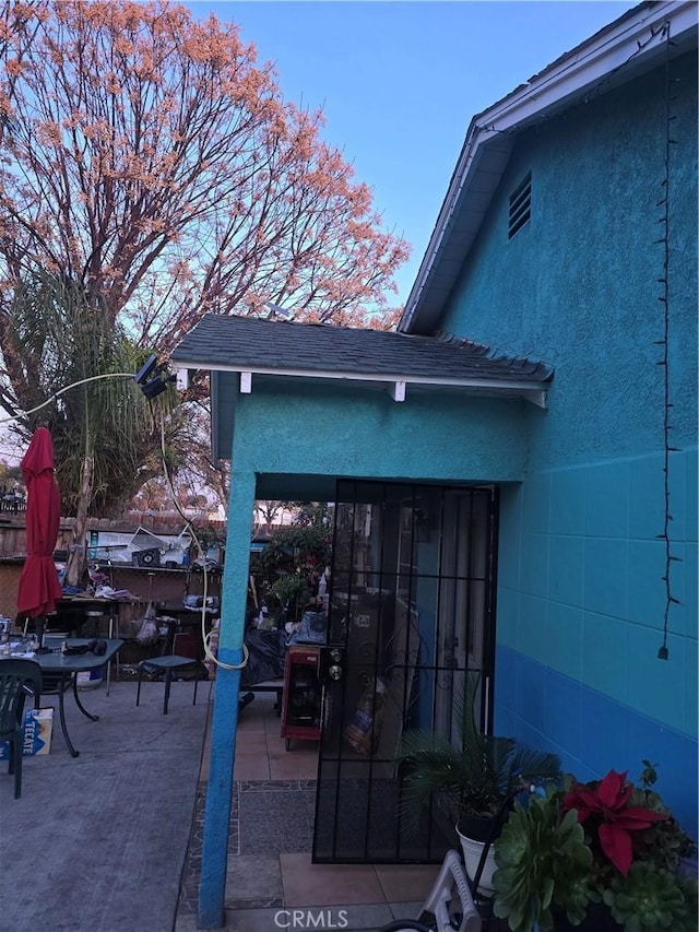 entrance to property featuring a shingled roof, a patio, and stucco siding