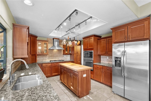 kitchen with wooden counters, appliances with stainless steel finishes, brown cabinetry, a sink, and wall chimney exhaust hood