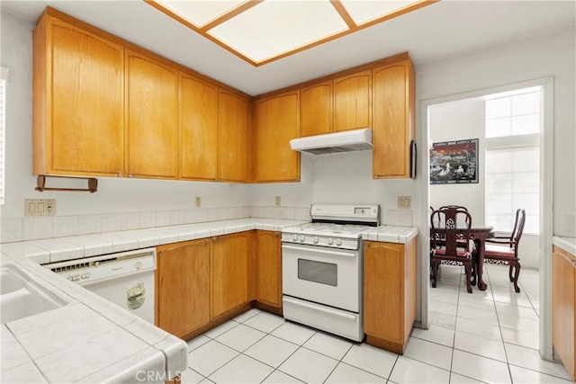 kitchen with white appliances, tile counters, under cabinet range hood, a sink, and light tile patterned flooring