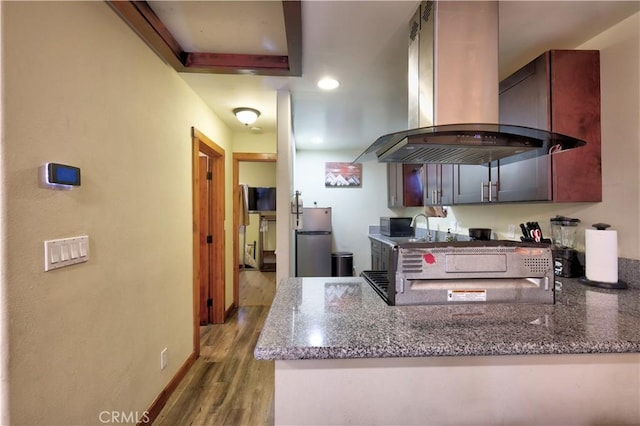 kitchen featuring a peninsula, wood finished floors, freestanding refrigerator, dark stone counters, and island exhaust hood