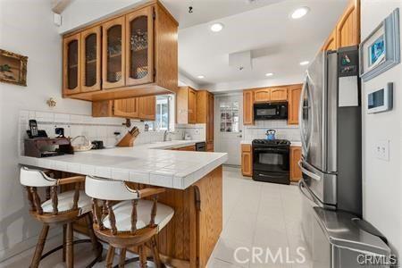 kitchen featuring tile counters, a kitchen bar, brown cabinetry, a peninsula, and black appliances