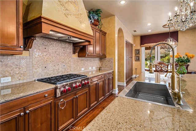 kitchen with light stone counters, arched walkways, stainless steel gas cooktop, an inviting chandelier, and premium range hood