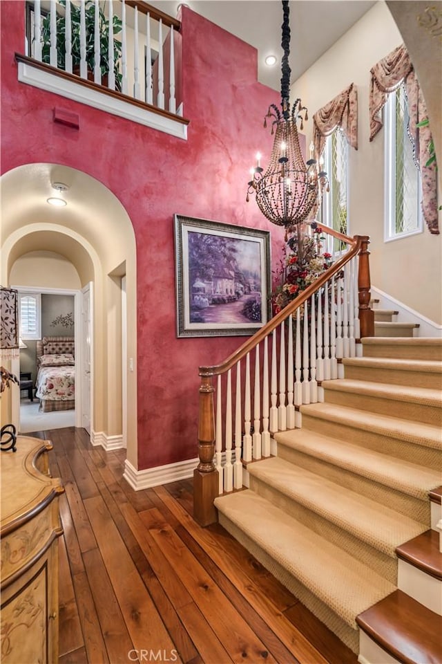 staircase featuring arched walkways, a notable chandelier, a high ceiling, wood finished floors, and baseboards