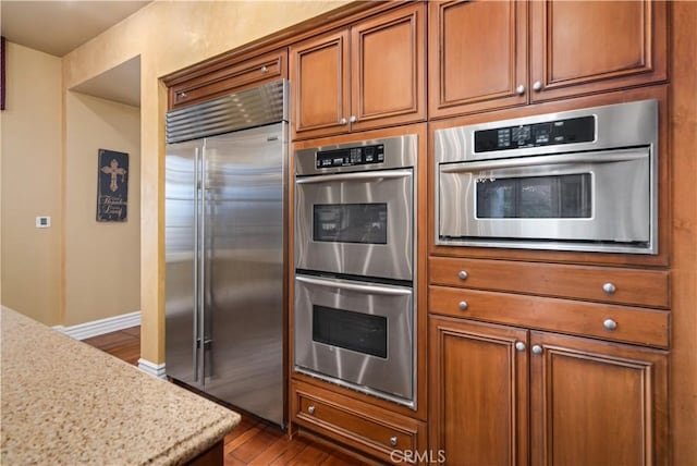 kitchen featuring baseboards, dark wood finished floors, brown cabinetry, light stone countertops, and stainless steel appliances