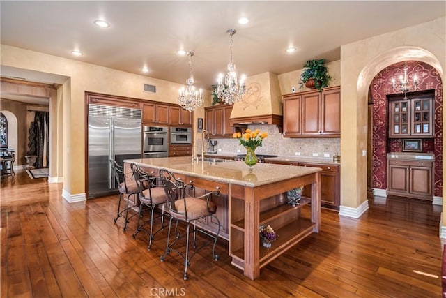 kitchen with appliances with stainless steel finishes, open shelves, an island with sink, decorative light fixtures, and custom range hood