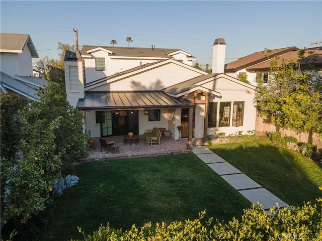 rear view of house with a patio, a chimney, a standing seam roof, fence, and a yard