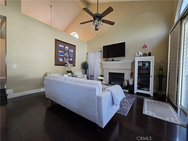 living room with dark wood-type flooring, a ceiling fan, a brick fireplace, high vaulted ceiling, and baseboards