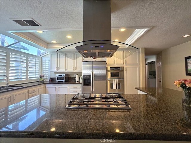 kitchen with island exhaust hood, stainless steel appliances, white cabinets, a sink, and dark stone countertops