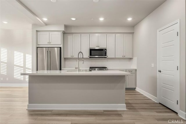 kitchen featuring light wood finished floors, light countertops, decorative backsplash, appliances with stainless steel finishes, and an island with sink