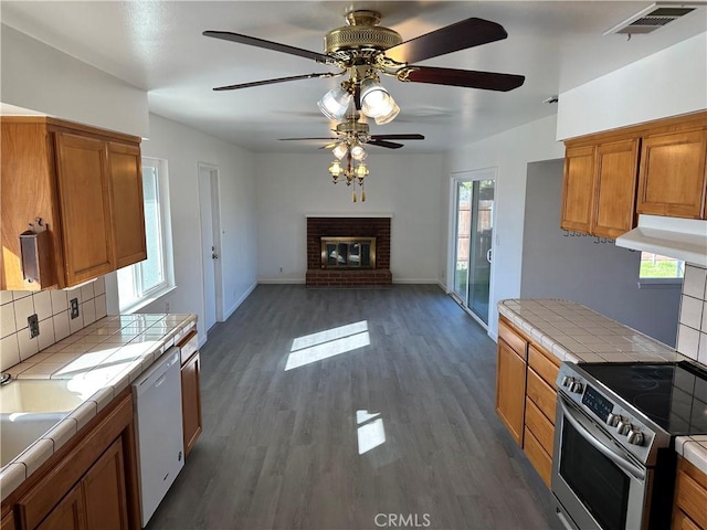 kitchen featuring electric stove, tile countertops, a brick fireplace, white dishwasher, and under cabinet range hood