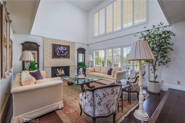 living area with a brick fireplace, dark wood-style flooring, a towering ceiling, and baseboards