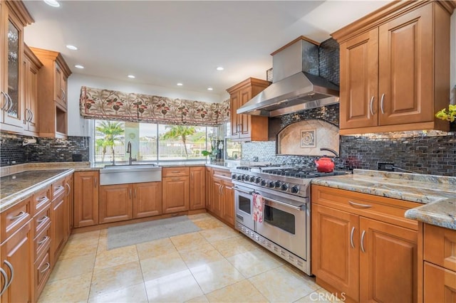 kitchen featuring wall chimney exhaust hood, glass insert cabinets, light stone countertops, double oven range, and a sink