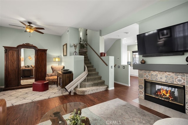 living area featuring ceiling fan, wood finished floors, baseboards, stairway, and a tiled fireplace